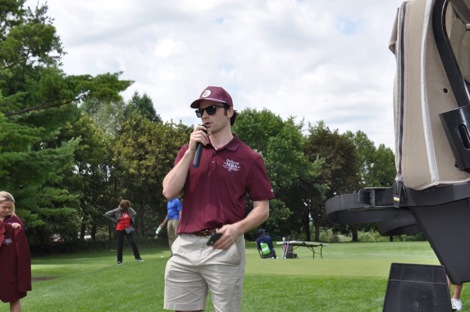 Committee member in a maroon polo on the microphone beside a cart